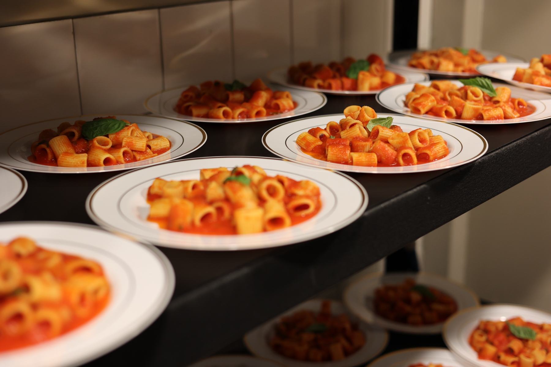 Rows of plated rigatoni on gold-rimmed plates, lined up on a service rack ready to go out.